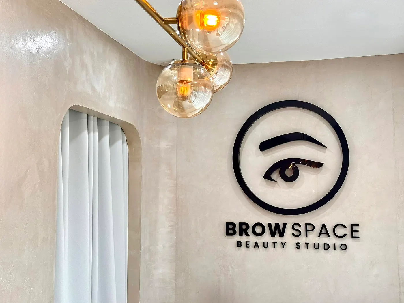Inside Brow Space: limewash wall, the black line-art logo, an arched doorway and gold pendant lights